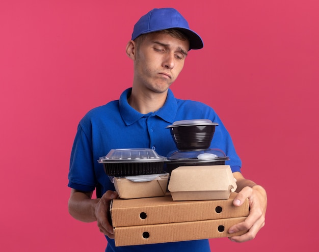 Un jeune livreur blond déçu tient et regarde des contenants de nourriture et des emballages sur des boîtes à pizza isolées sur un mur rose avec espace de copie