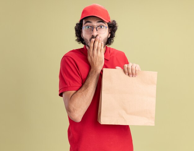 Jeune livreur caucasien concerné en uniforme rouge et casquette portant des lunettes tenant un paquet de papier gardant la main sur la bouche isolée sur un mur vert olive avec espace de copie