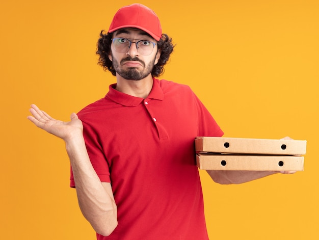 Jeune livreur caucasien désemparé en uniforme rouge et casquette portant des lunettes tenant des paquets de pizza montrant une main vide isolée sur un mur orange