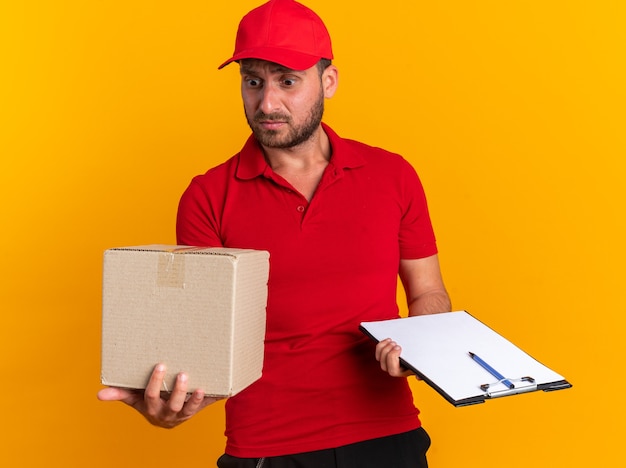 Jeune livreur caucasien désemparé en uniforme rouge et casquette tenant un presse-papiers avec un stylo et une boîte en carton regardant une boîte en carton isolée sur un mur orange