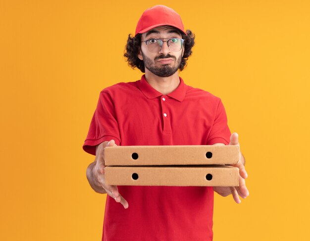 Photo gratuite jeune livreur caucasien impressionné en uniforme rouge et casquette avec des paquets de pizza