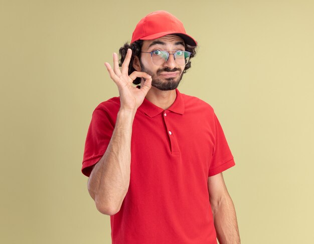 Jeune livreur caucasien impressionné en uniforme rouge et casquette portant des lunettes faisant un geste délicieux