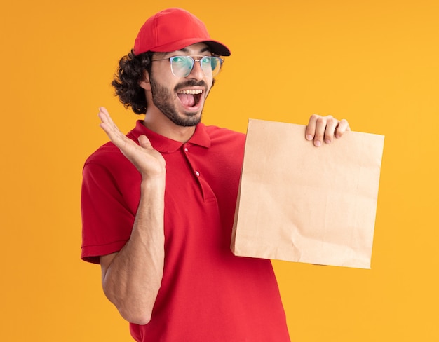 Jeune livreur caucasien impressionné en uniforme rouge et casquette portant des lunettes tenant un paquet de papier montrant la main vide