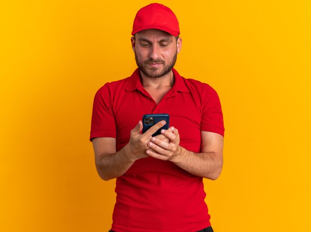 Jeune livreur caucasien sérieux en uniforme rouge et casquette tenant et regardant un téléphone portable isolé sur un mur orange avec espace pour copie