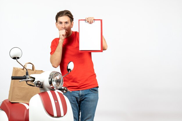 Jeune livreur choqué et émotionnel en uniforme rouge debout près de scooter montrant un document sur un mur blanc