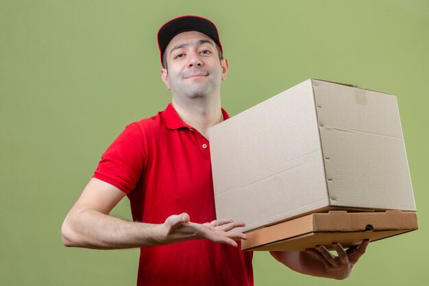 Jeune livreur confiant portant uniforme rouge tenant des boîtes en carton avec sourire sur le visage sur mur vert isolé