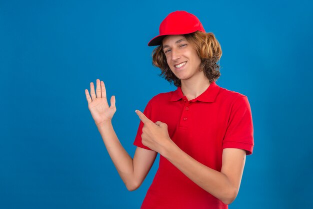 Jeune livreur confiant en uniforme rouge souriant confiant pointant avec la main et le doigt sur le côté sur mur bleu isolé