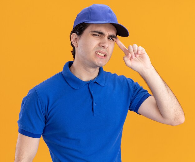 Jeune livreur confus en uniforme bleu et casquette regardant devant faisant un geste de réflexion isolé sur un mur orange