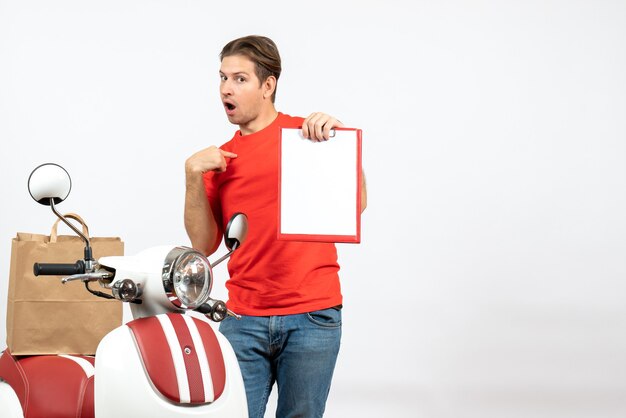 Jeune livreur confus en uniforme rouge debout près de scooter montrant le document se pointant sur le mur blanc