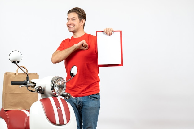 Jeune livreur fier en uniforme rouge debout près de scooter montrant un document sur un mur blanc