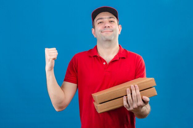 Jeune livreur heureux en polo rouge et casquette tenant des boîtes à pizza à la confiance en levant le poing après un concept gagnant de la victoire debout sur fond bleu isolé