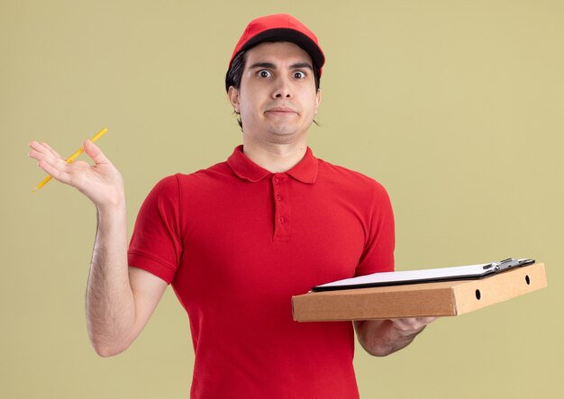 Jeune livreur impressionné en uniforme rouge et casquette tenant le presse-papiers et le crayon de paquet de pizza regardant l'avant isolé sur le mur vert olive