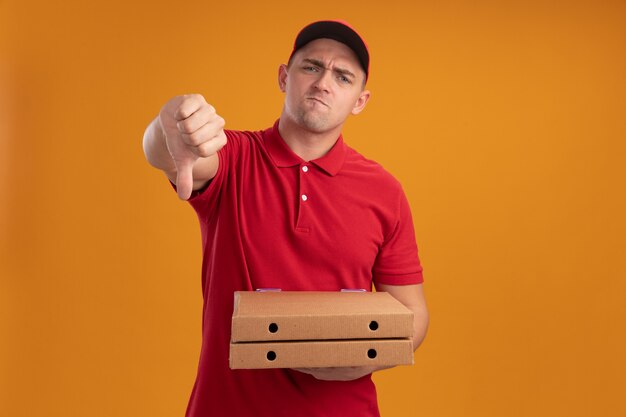 Jeune livreur insatisfait en uniforme avec capuchon tenant des boîtes de pizza montrant le pouce vers le bas isolé sur un mur orange