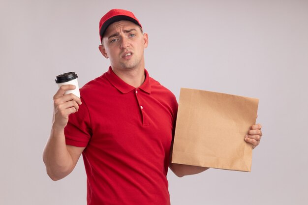 Jeune livreur insatisfait en uniforme avec capuchon tenant un paquet de papier alimentaire avec une tasse de café isolé sur un mur blanc