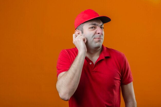 Jeune livreur mécontent en polo rouge et casquette se gratter l'oreille n'ayant aucune idée de quoi faire ensuite sur fond orange isolé