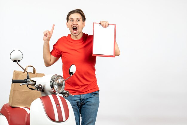 Jeune livreur nerveux et émotionnel en uniforme rouge debout près de scooter montrant un document sur un mur blanc