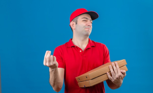 Jeune livreur en polo rouge et cap tenant des boîtes de pizza faisant de délicieux geste italien avec main souriant sur mur bleu isolé