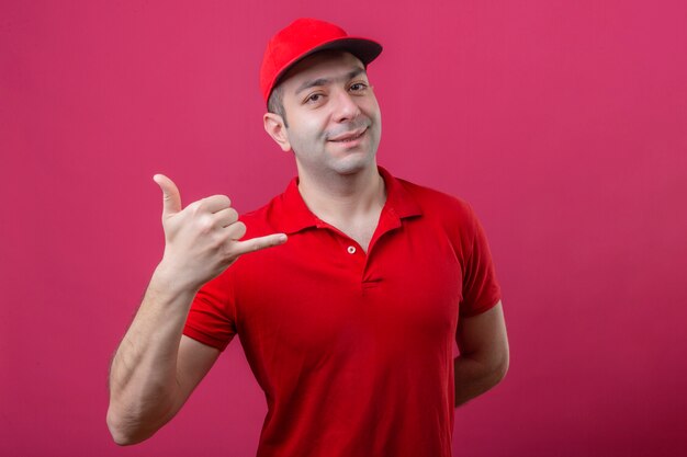 Jeune livreur en polo rouge et casquette faisant appelez-moi geste à la confiance souriant joyeusement sur fond rose isolé