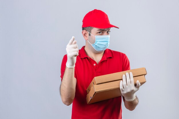 Jeune livreur en polo rouge et casquette en masque médical tenant des boîtes de pizza à la caméra avec reproche et pointant le doigt sur vous sur mur blanc isolé