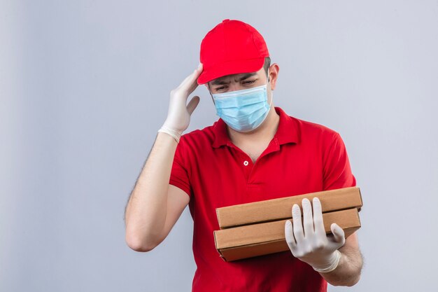 Jeune livreur en polo rouge et une casquette en masque médical tenant des boîtes à pizza à la tête malade souffrant de maux de tête sur mur blanc isolé