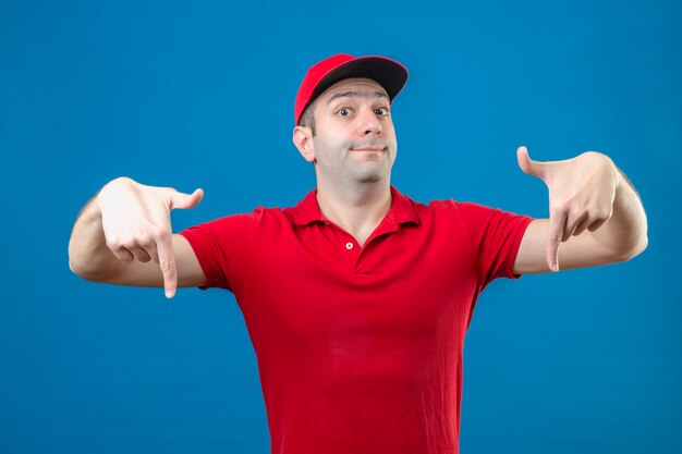 Jeune livreur en polo rouge et casquette pointant avec le doigt vers le bas en regardant la caméra avec sourire expression satisfaite sur fond bleu isolé