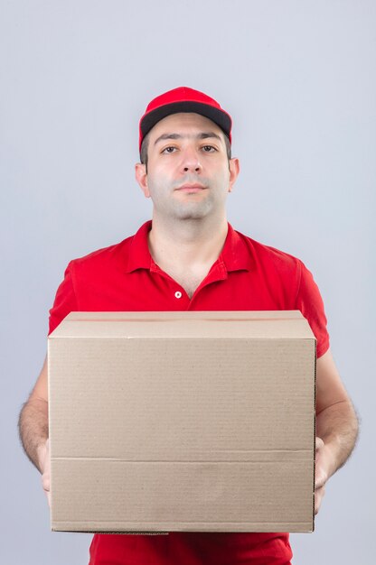 Jeune livreur en polo rouge et casquette tenant une boîte en carton avec un visage sérieux debout sur un mur blanc isolé