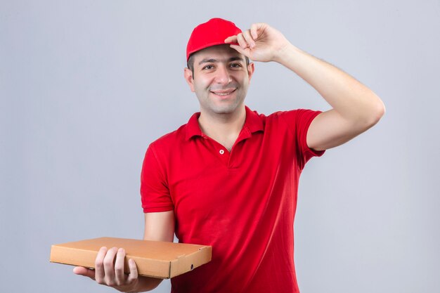 Jeune livreur en polo rouge et casquette tenant des boîtes à pizza faisant un geste de salutation touchant sa casquette smiling friendly sur mur blanc isolé