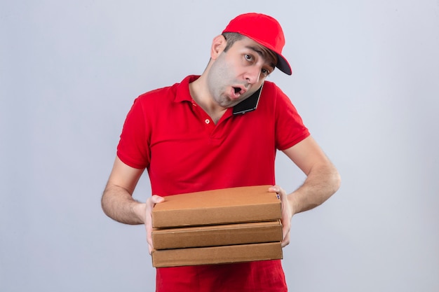 Jeune livreur en polo rouge et casquette tenant des boîtes à pizza tout en parlant au téléphone mobile à la surprise sur mur blanc isolé