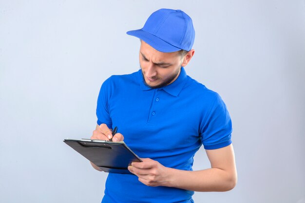 Jeune livreur portant un polo bleu et une casquette debout avec presse-papiers et écrit avec un visage sérieux sur fond blanc isolé