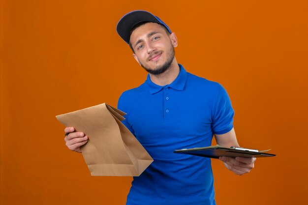 Jeune livreur portant un polo bleu et une casquette debout avec presse-papiers et paquet de papier regardant la caméra avec le sourire sur le visage sur fond orange isolé