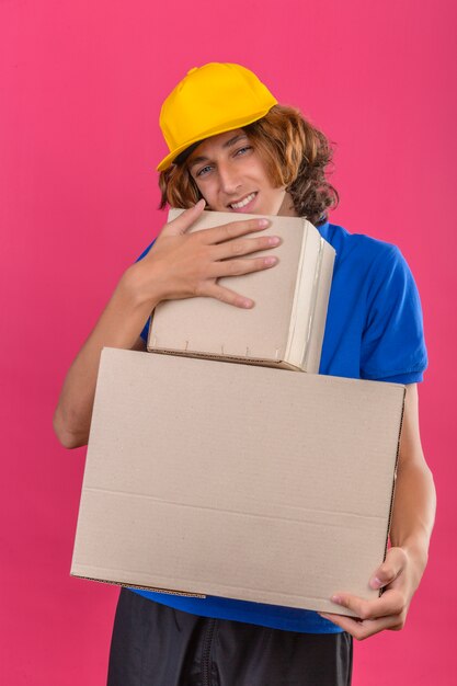 Jeune livreur portant un polo bleu et une casquette jaune tenant des boîtes en carton rêvant d'embrasser des boîtes souriant avec un visage heureux sur fond rose isolé
