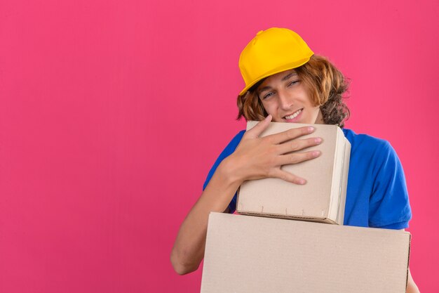 Photo gratuite jeune livreur portant un polo bleu et une casquette jaune tenant des boîtes en carton rêvant d'embrasser des boîtes souriant avec un visage heureux sur fond rose isolé