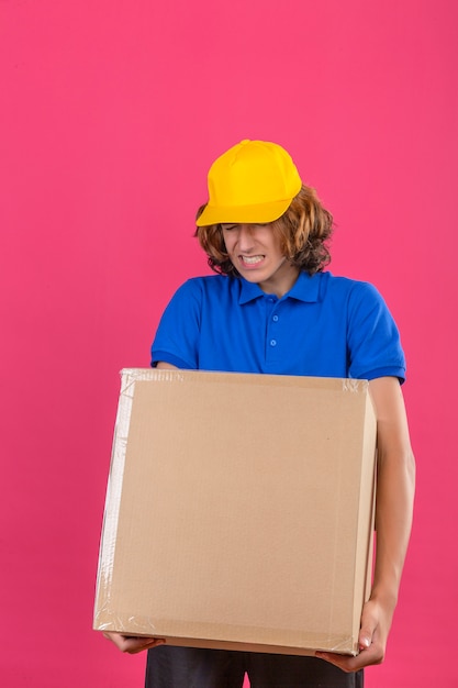 Photo gratuite jeune livreur portant un polo bleu et une casquette jaune tenant une grosse grosse boîte en carton lourd se sentir mal à cause du poids lourd sur fond rose isolé