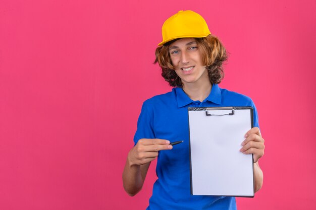 Jeune livreur portant un polo bleu et une casquette jaune tenant le presse-papiers et un stylo dans les mains demandant la signature à la caméra en souriant sympathique sur fond rose isolé