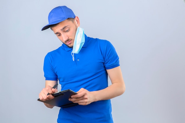 Jeune livreur portant un polo bleu et une casquette avec mas de protection médicale sur l'oreille en prenant des notes dans le presse-papiers sur fond blanc isolé