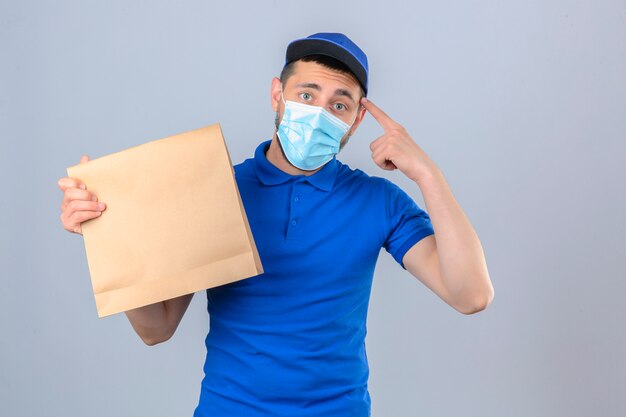 Jeune livreur portant un polo bleu et une casquette en masque médical de protection debout avec un paquet de papier pointant vers la tête sur fond blanc isolé