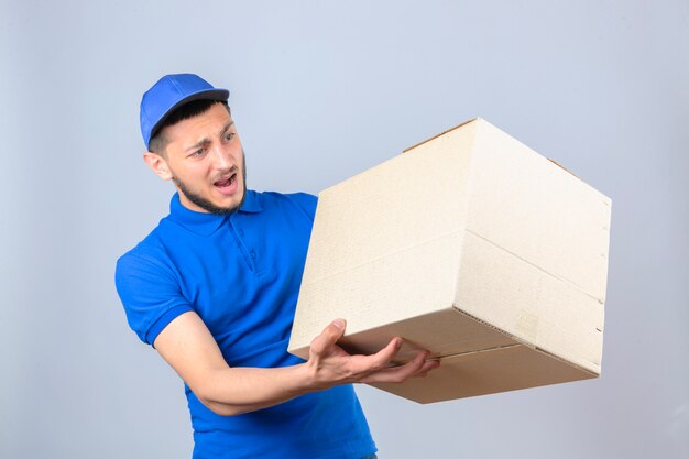 Jeune livreur portant un polo bleu et une casquette à la surprise et confus debout avec boîte en carton sur fond blanc isolé