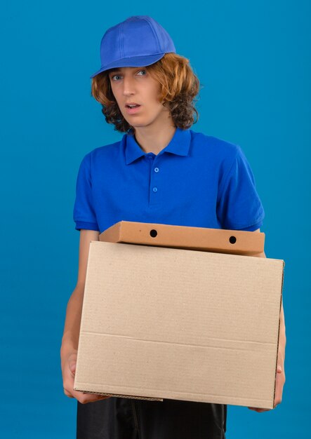 Jeune livreur portant un polo bleu et une casquette tenant des boîtes en carton dans les mains à la surprise debout sur fond bleu isolé