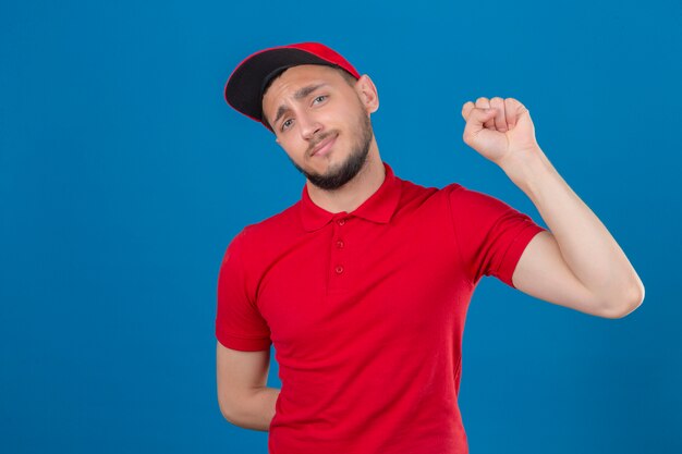 Jeune livreur portant un polo rouge et une casquette en levant le poing après un concept gagnant de la victoire sur fond bleu isolé