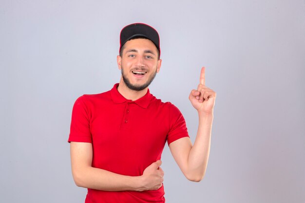 Jeune livreur portant un polo rouge et une casquette souriant joyeusement pointant vers le haut avec le doigt sur fond blanc isolé