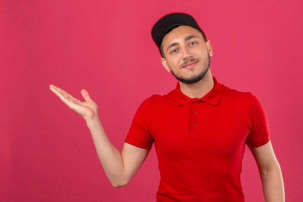 Jeune livreur portant un polo rouge et une casquette souriant présentant joyeusement et pointant avec la paume de la main en regardant la caméra sur fond rose isolé