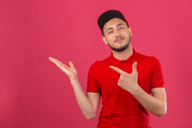 Jeune livreur portant un polo rouge et une casquette souriant et regardant la caméra pointant avec deux mains et doigts sur le côté sur fond rose isolé