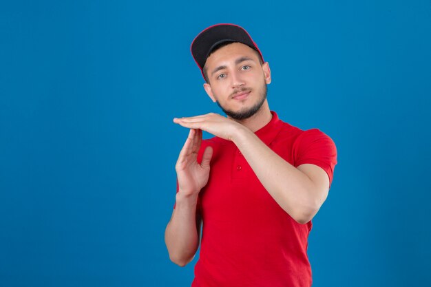 Jeune livreur portant un polo rouge et une casquette à la surmenage faisant le geste de temps avec les mains sur fond bleu isolé