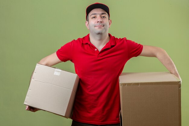 Jeune livreur portant l'uniforme rouge à la confiance debout avec de grandes boîtes en carton dans les mains sur fond vert isolé