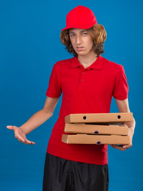 Jeune livreur sceptique en uniforme rouge tenant des boîtes de pizza se sentir confus sur fond bleu isolé
