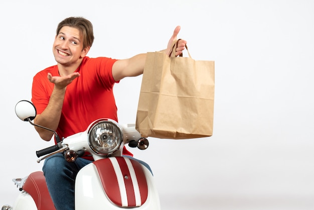 Photo gratuite jeune livreur souriant en uniforme rouge assis sur un scooter donnant un sac en papier à quelqu'un sur le mur jaune