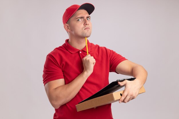 Jeune livreur stricte en uniforme avec capuchon tenant le presse-papiers avec boîte à pizza et mettre un crayon sur le menton isolé sur un mur blanc