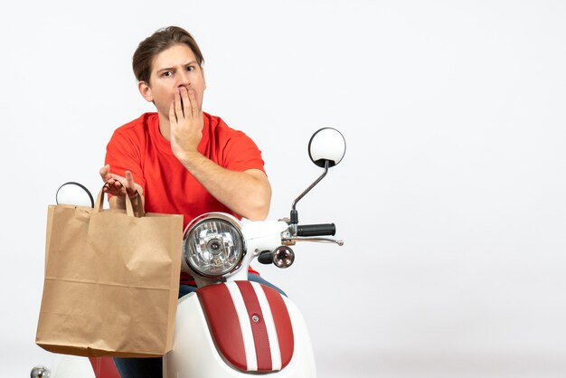 Jeune livreur surpris en uniforme rouge assis sur un scooter tenant un sac en papier sur un mur blanc