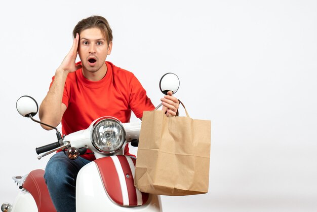 Jeune livreur surpris en uniforme rouge assis sur un scooter tenant un sac en papier posant pour l'avant sur un mur blanc