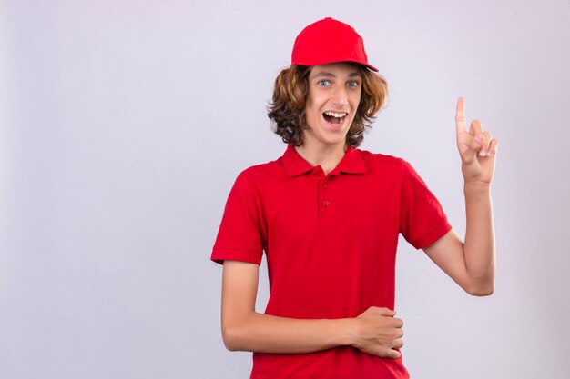 Jeune livreur surpris en uniforme rouge pointant vers le haut une excellente idée souriant avec visage heureux sur fond isolé